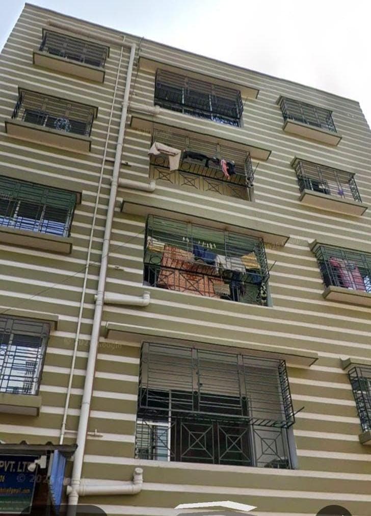 Chaitali Co-operative Housing Society from Street No. 152 — four floors of grilled windows and shallow balconies on a banded cream facade, a rainwater downpipe running the height of it, and the company signboard at ground level